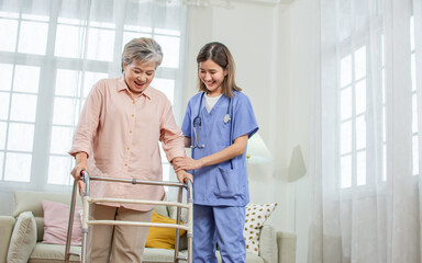 Asian beautiful female nurse or doctor wearing uniform, helping 60s elderly woman to walk by walker, assisting, smiling with happiness, standing in nursing home. Retirement, Healthcare Concept.