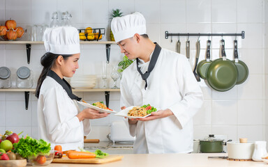 Two Asian male and female professional chef wearing uniform, holding plate, presenting of cooking competition, looking each other, standing in kitchen.