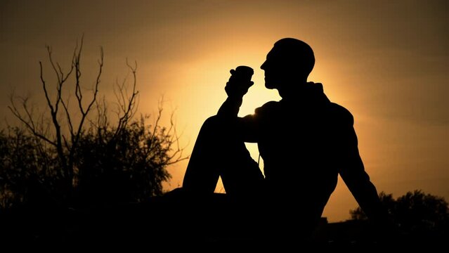 Silhouetted man at sunset drinks coffee on a bench. Fragrant coffee in the evening in nature. A guy enjoys coffee at sunset. Thoughts alone with nature. Free thinking concept.