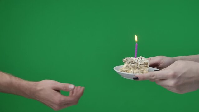 Close up on green chromakey background. A woman and a man, you can see their hands. She holds a piece of cake with a burning candle and hands it to the man. It's his birthday, she congratulates him