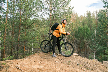 An active lifestyle.A cyclist with a backpack in a protective helmet stands on a mountain with a mountain bike in autumn among the trees, side view