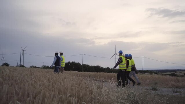 Side view group engineers in wind power plant walking together at sunset for maintenance windmill wear safety clothes and equipped with working tools. Professional technicians men in charge of repair.