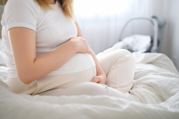 A soon to be mother, dressed in light attire, sits on the bed and lovingly cradles her pregnant belly.