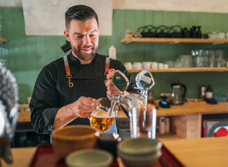 Smiling stylish bearded barman dressed black uniform with an apron tapping fresh lager beer into glass mug at bar counter. Successful people, beer consumption, beverages industry concept image