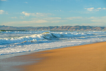 Light airy seascape. Wide sandy beach, sea waves, silhouette of mountains, and cloudy sky