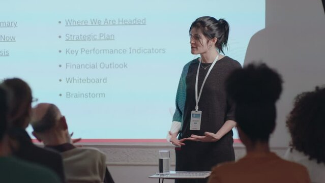 Young female speaker walking into conference hall, standing in front of projection screen and giving business presentation about strategic plan to audience during corporate seminar