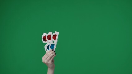 In the close up shot on the green background. A womans hand that is raised up and holds 3D glasses...