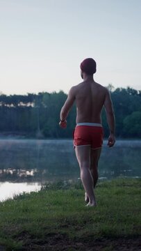 Good looking man in swim shorts going to the lake through the grass and looking at the water