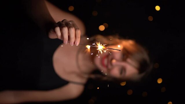 Happy smiling blonde woman holding Bengal light sparkler. Christmas lights at black background. Slow motion vertical video footage. Celebration birthday or New year party time 