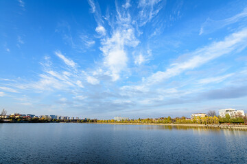 Landscape with trees and the lake in Linden Park (Parcul Tei), in Bucharest, Romania, in a sunny autumn day with white clouds and blue sky - obrazy, fototapety, plakaty