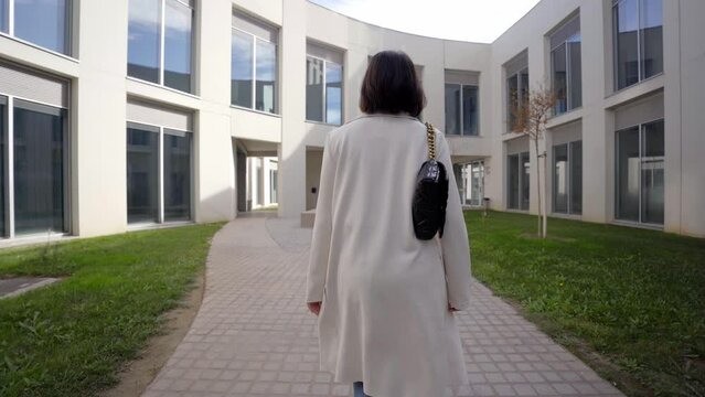 Camera follows business woman in formal suit going to work through a path of modern buildings. Elegant girl with trench coat and handbag on her way to office. Concept of success and career. Rear view