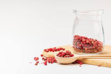 Pink peppercorns in a wooden spoon and glass jar on wooden stand on a white background with copy space