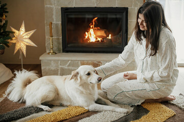 Beautiful woman in stylish pajamas relaxing with cute white dog at cozy fireplace, enjoying calm...