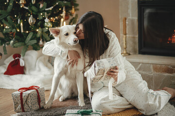 Cozy christmas morning. Beautiful woman in pajamas holding stylish christmas gift and hugging cute...