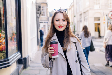 Young smiling woman in trench coat enjoying hot coffee drink in reusable cup while walking on the city streets decorated for the autumn holidays. Happy fall cozy mood, seasonal fashion style trend. - obrazy, fototapety, plakaty