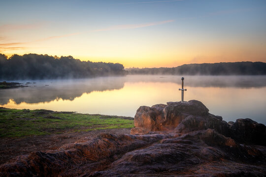 Excalibur l'épée légendaire du roi Arthur,  plantée sur son rocher devant un étang dans la brume du matin au lever du soleil en forêt de Brocéliande
