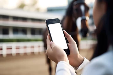 Poster Closeup of mobile phone with white blank screen in hands of girl in stands at horse racing outdoors. © Bonsales