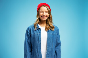 Portrait of beautiful smiling woman wearing red hipster hat and stylish jeans jacket isolated on blue background. Female tourist with wavy silky hair looking at camera. Travel concept