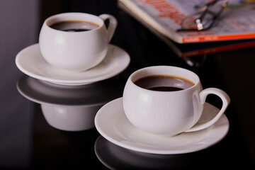 Cups of fresh coffee on a black coffee table with a magazine and some reading glasses in the background.