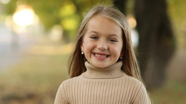 Adorable cheerful little child girl 5-6 years old smiles with teeth at camera outdoors on autumn warm day. Preschool kid with long hair wears beige knit longsleeve. Children's day. Happy childhood