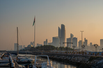 Fototapeta na wymiar Abu Dhabi, UAE, skyline from across the Corniche with a harbour, at sunrise. Tall UAE flag in the foreground.