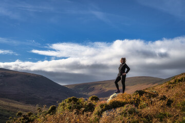 Adult woman standing on the rock, illuminated by sunlight, and looking at distant mountains. Hiking in Wicklow Mountains, Ireland