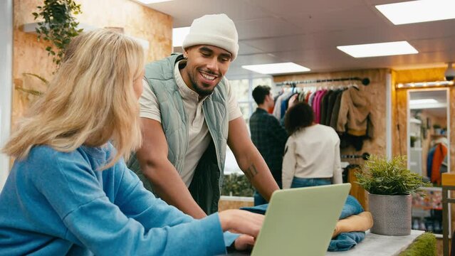 Male and female sales assistants working at cash desk with laptop in pop up fashion or clothes store with customers in background - shot in slow motion