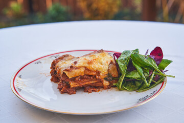Delicious tasty and juicy homemade lasagne in Naples style served on the porcelain plate with fresh green lettuce salad outside on the table in the home garden. Traditional recipe from south Italy.