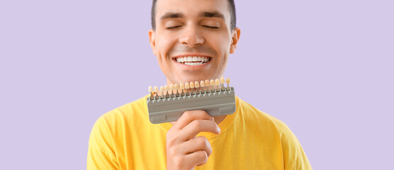 Happy young man with teeth color samples on lilac background