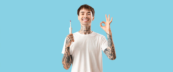 Young tattooed man with electric toothbrush showing OK on light blue background