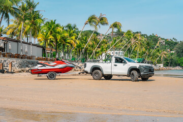 White pickup with jetski on trailer stands on sand beach, close to restaurant terrace and palm trees, morning time, tropical resort, Thailand - obrazy, fototapety, plakaty