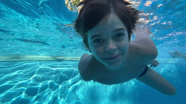 Slow motion video of happy boy of 9 years old diving and playing in the water of a swimming pool in the summer, blue water, smiling, and funny way.