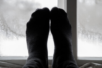 Feet in gray socks on a warm windowsill against a frosty window in a cold winter. Warm homely mood.
