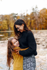 Fototapeta premium Very charming beautiful brunette mother hugs little girl, her daughter and smiles happily against the background of yellow leaves and lake in the park in nature