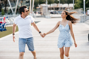 A couple in love, a man and a woman, walk along a wooden pier. A man and a woman hold hands and run along a wooden pier. Stylish man and his beautiful woman smiling and having fun