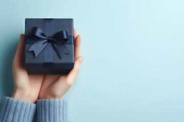 Top view to young woman hands holding navy blue present gift giftbox with satin ribbon bow on light blue background. Christmas, birthday, anniversary, shopping, blue friday concept