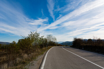 Asphalt road a top a mountain, framed by a vivid blue sky streaked with picturesque white clouds. The empty, newly laid two lane road leads toward distant hills and mountains