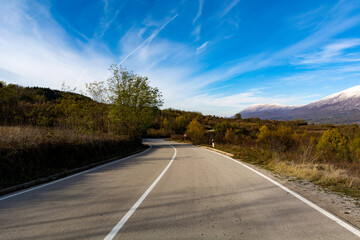 Autumns sunlit asphalt road atop a mountain, flanked by foliage and trees with golden yellow leaves. In the distance, a snow capped mountain peak under the sunny autumn sky