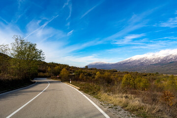 Meandering asphalt road winding through nature on a hill on an autumn day. A snow capped mountain peak is visible in the distance under the beautiful blue sky adorned with white clouds