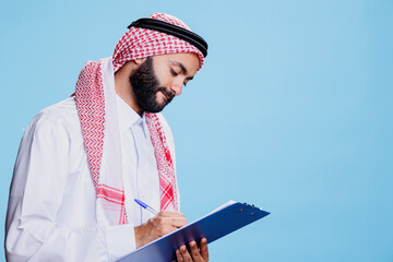 Man wearing traditional islamic thobe and headscarf clothes writing in clipboard, filling...