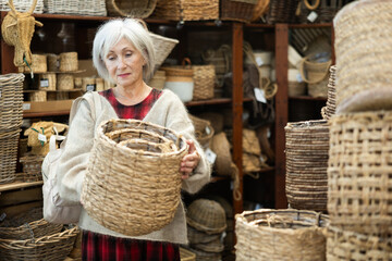 Elderly woman shopper choosing wicker basket in store..
