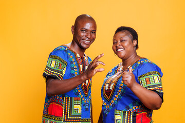 Fototapeta na wymiar Smiling african american man and woman wearing ethnic clothes showing thumb up and okay gesture portrait. Couple giving positive feedback and looking at camera on studio background