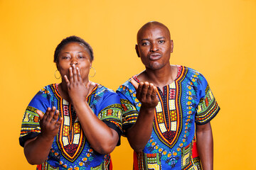 Obraz premium African american man and woman couple sending air kiss together, sharing love and looking at camera. Romantic boyfriend and girlfriend pair showing affection studio portrait