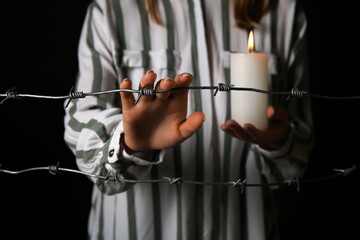 Jewish female prisoner with burning candle behind barbed wire on dark background, closeup....
