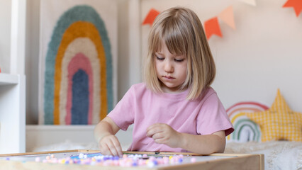 A cute girl is sitting at a table in the children's room and making crafts out of pompoms. Children's crafts.