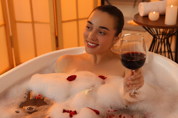 Happy woman with glass of wine taking bath in tub with foam and rose petals indoors