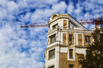 An old beige house, apartment building under construction on a city street. Construction crane machine over a roof and a blue cloudy sky. Restoration of an old building with stucco Roof reconstruction
