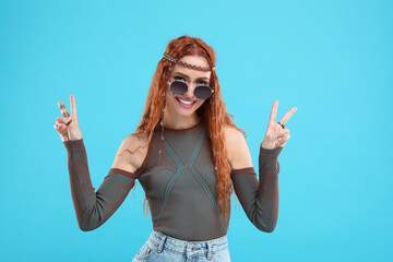 Stylish young hippie woman in sunglasses showing V-sign on light blue background
