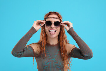 Stylish young hippie woman in sunglasses showing her tongue on light blue background - obrazy, fototapety, plakaty