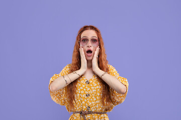 Surprised young hippie woman in sunglasses on violet background